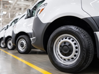 New white vans parked in a row inside factory, focus on the tire of the first van, yellow line on the floor.